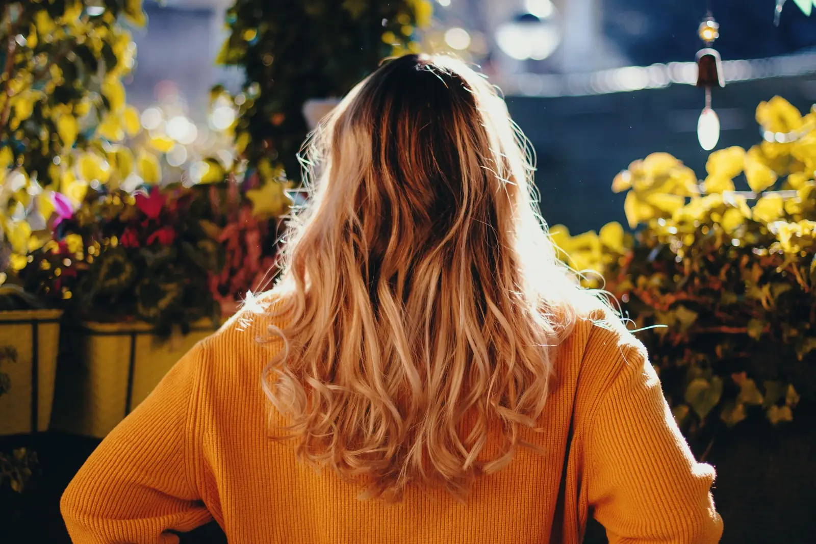 Mulher de costas, com cabelo loiro ondulado e suéter amarelo, diante de plantas iluminadas