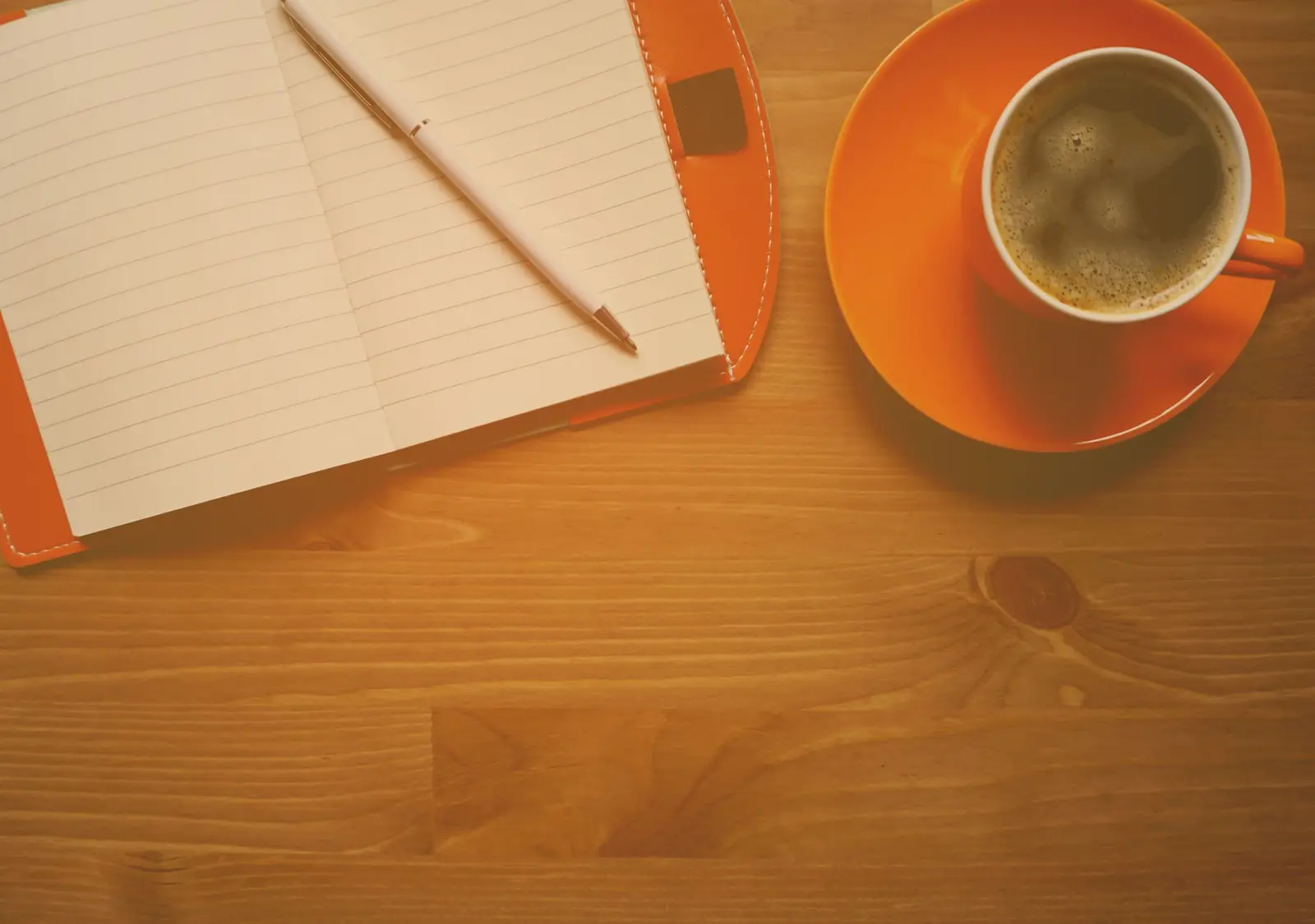 Caderno aberto com caneta ao lado de uma xícara de café laranja sobre mesa de madeira