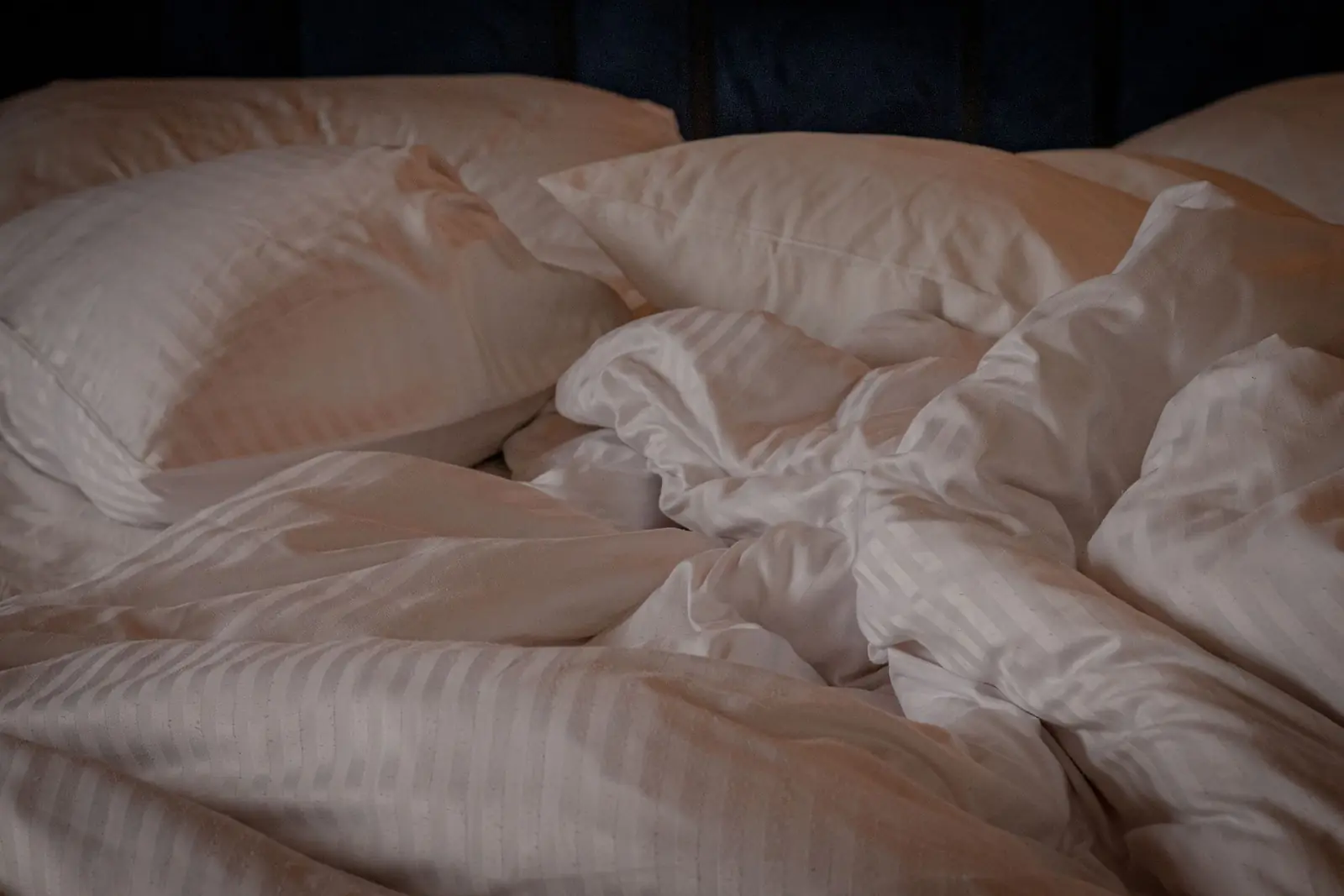 Cama desfeita na penumbra, com travesseiros e lençóis brancos amarrotados