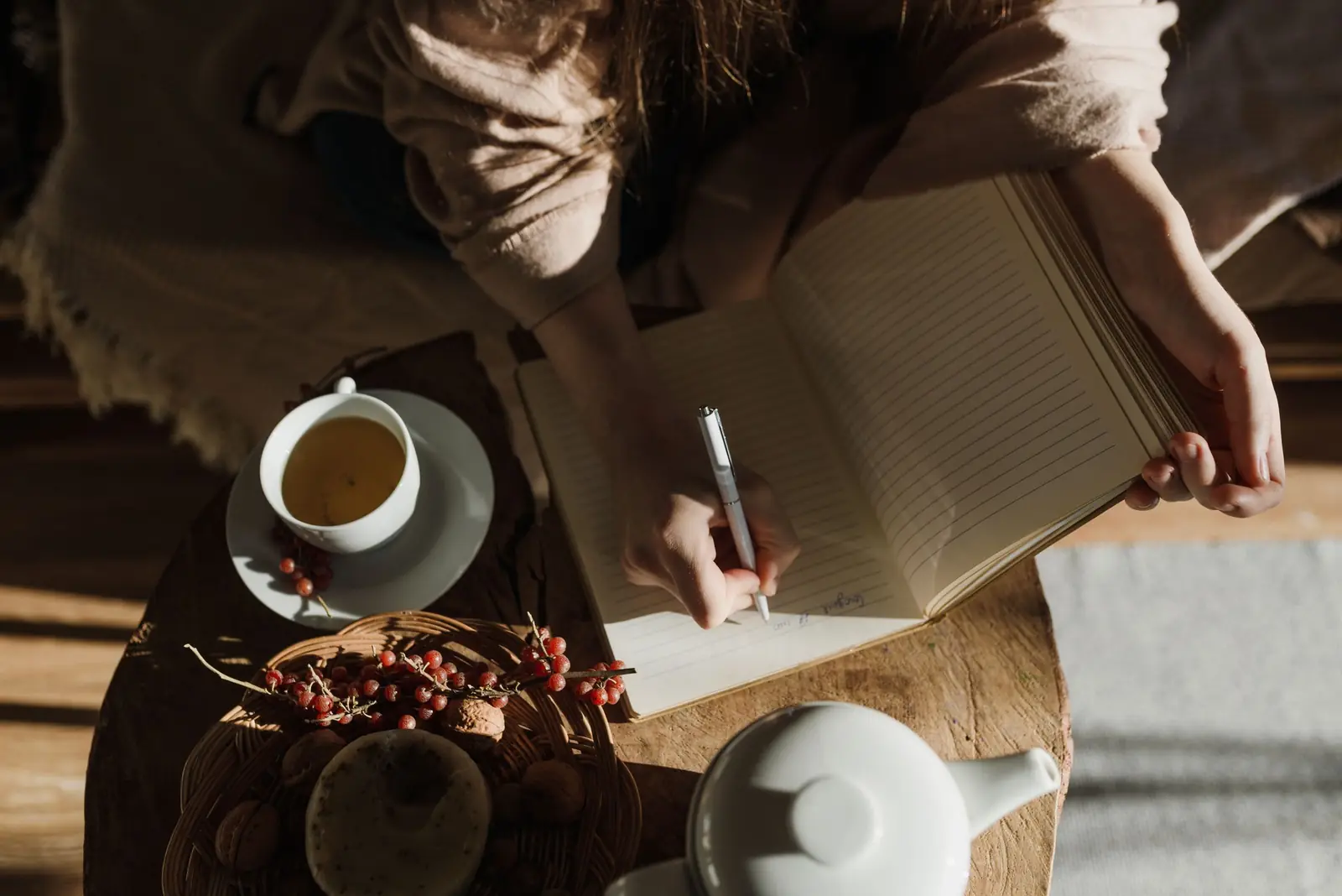 Mulher escrevendo em um caderno sobre mesa de madeira, ao lado de uma xícara de chá e um bule, à luz natural