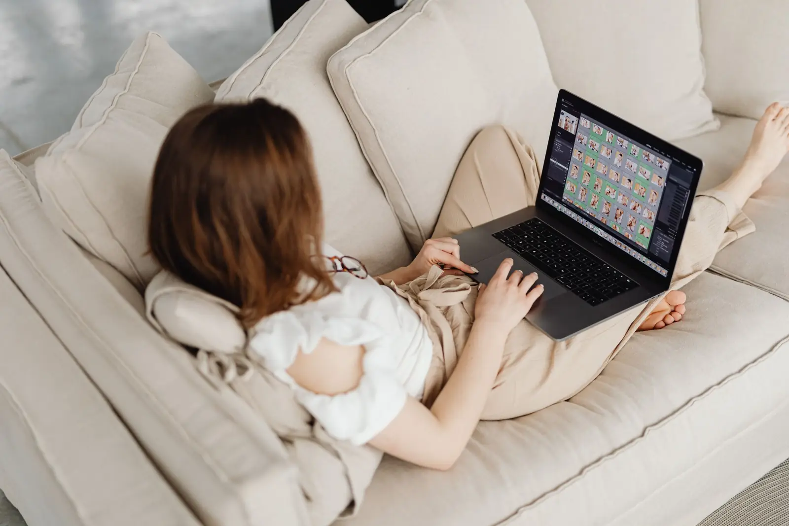 Mulher de costas, recostada em um sofá claro, usando um notebook