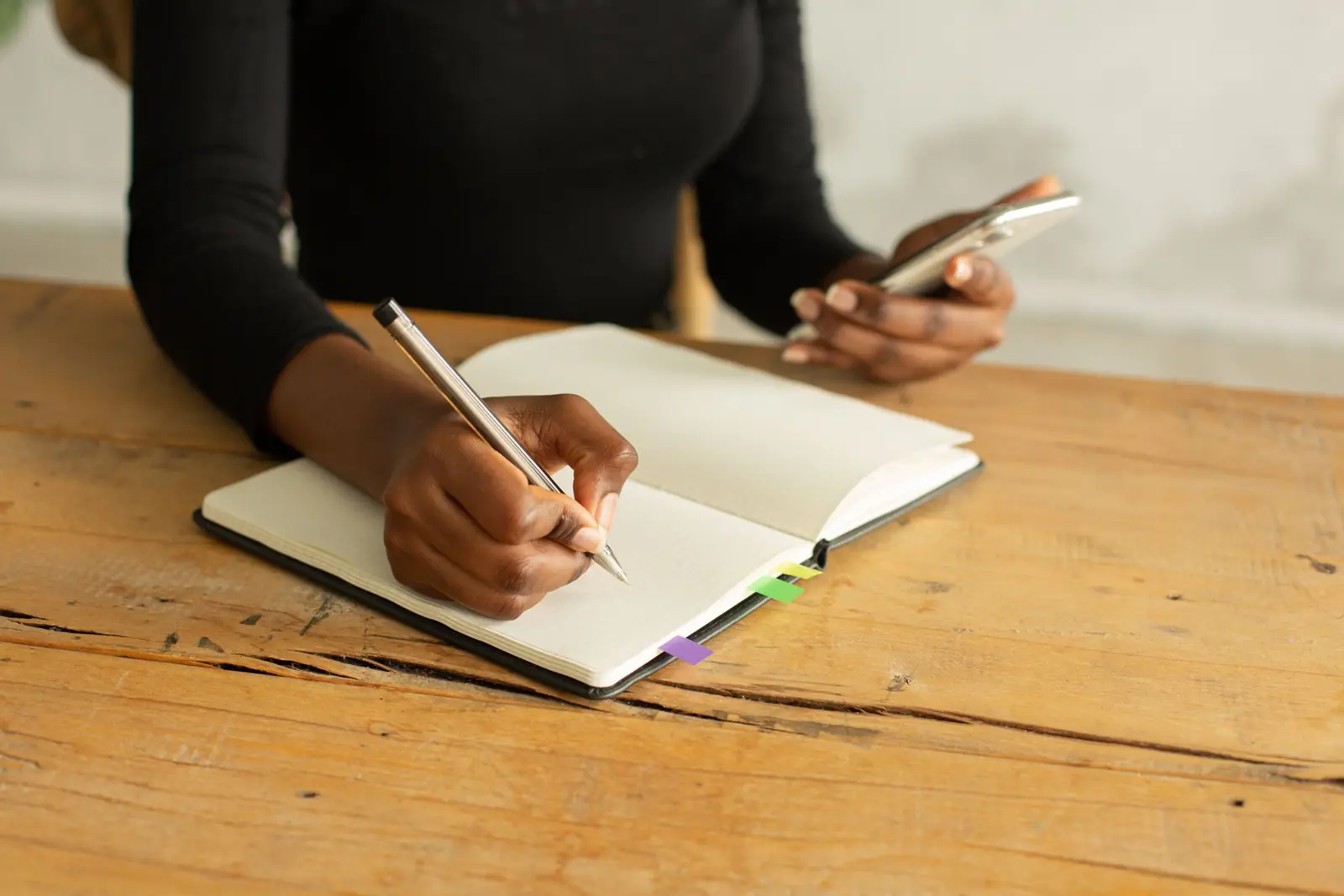 Mulher anota em um caderno enquanto segura o celular, sobre uma mesa de madeira