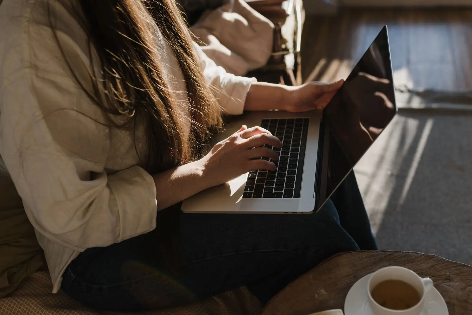 Mulher de cabelos longos usando o notebook no colo em casa, sob luz do sol, com uma xícara de chá ao lado