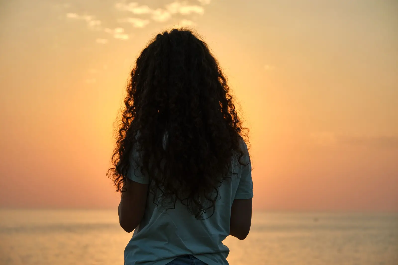 Mulher de cabelo cacheado, vista de costas, diante do mar ao pôr do sol
