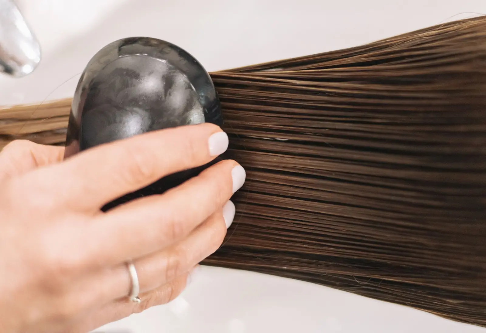 Mão segurando uma escova preta sobre mechas longas de cabelo castanho