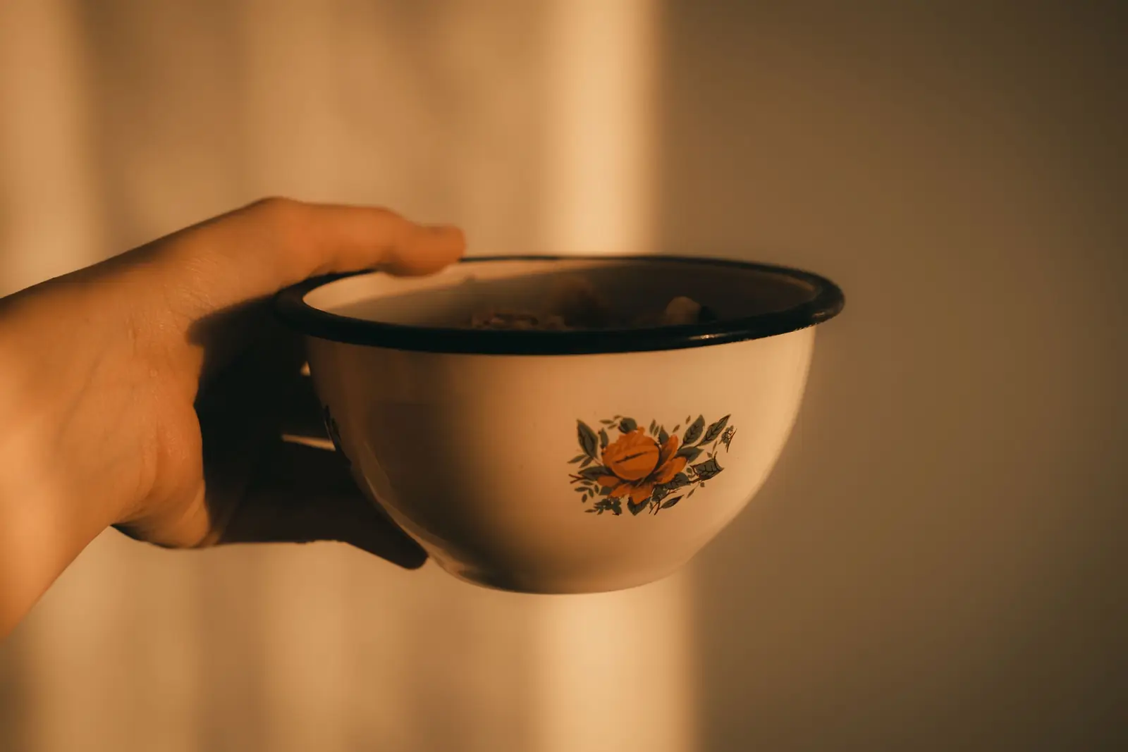Mão segura uma tigela esmaltada branca com estampa de flor laranja, sob luz quente de fim de tarde.