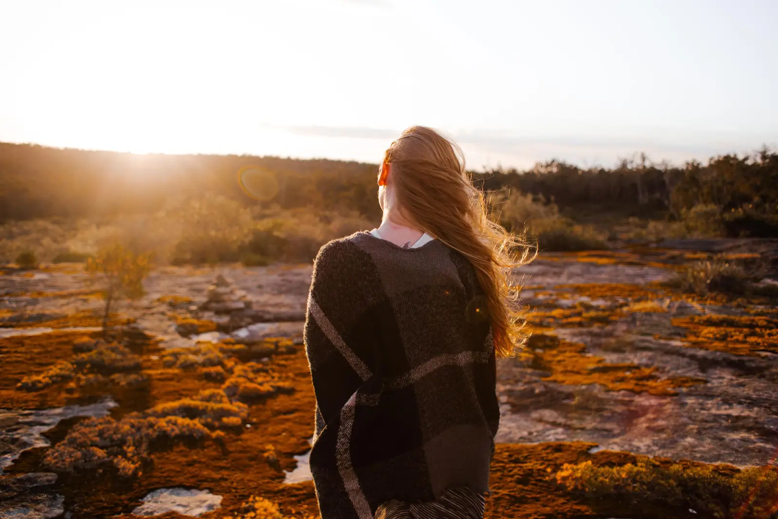 Mulher de costas, com cabelo ruivo ao vento, em um campo ao nascer do sol
