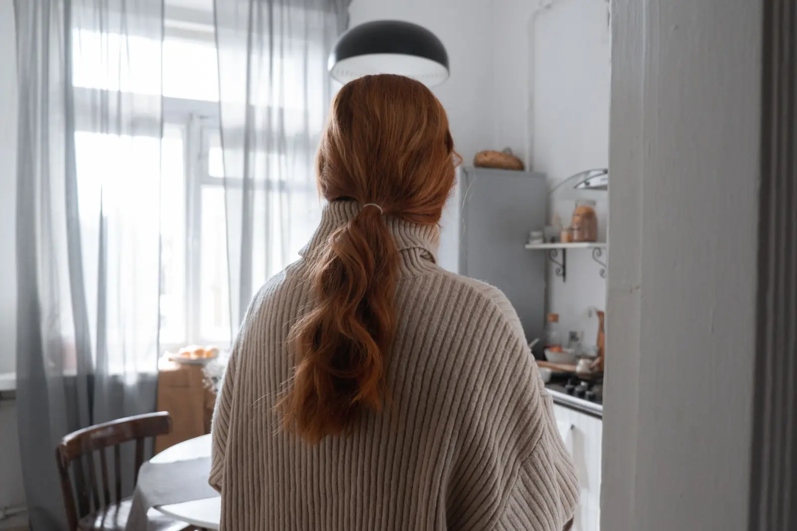 Mulher de costas, com rabo de cavalo ruivo e suéter bege, em uma cozinha clara