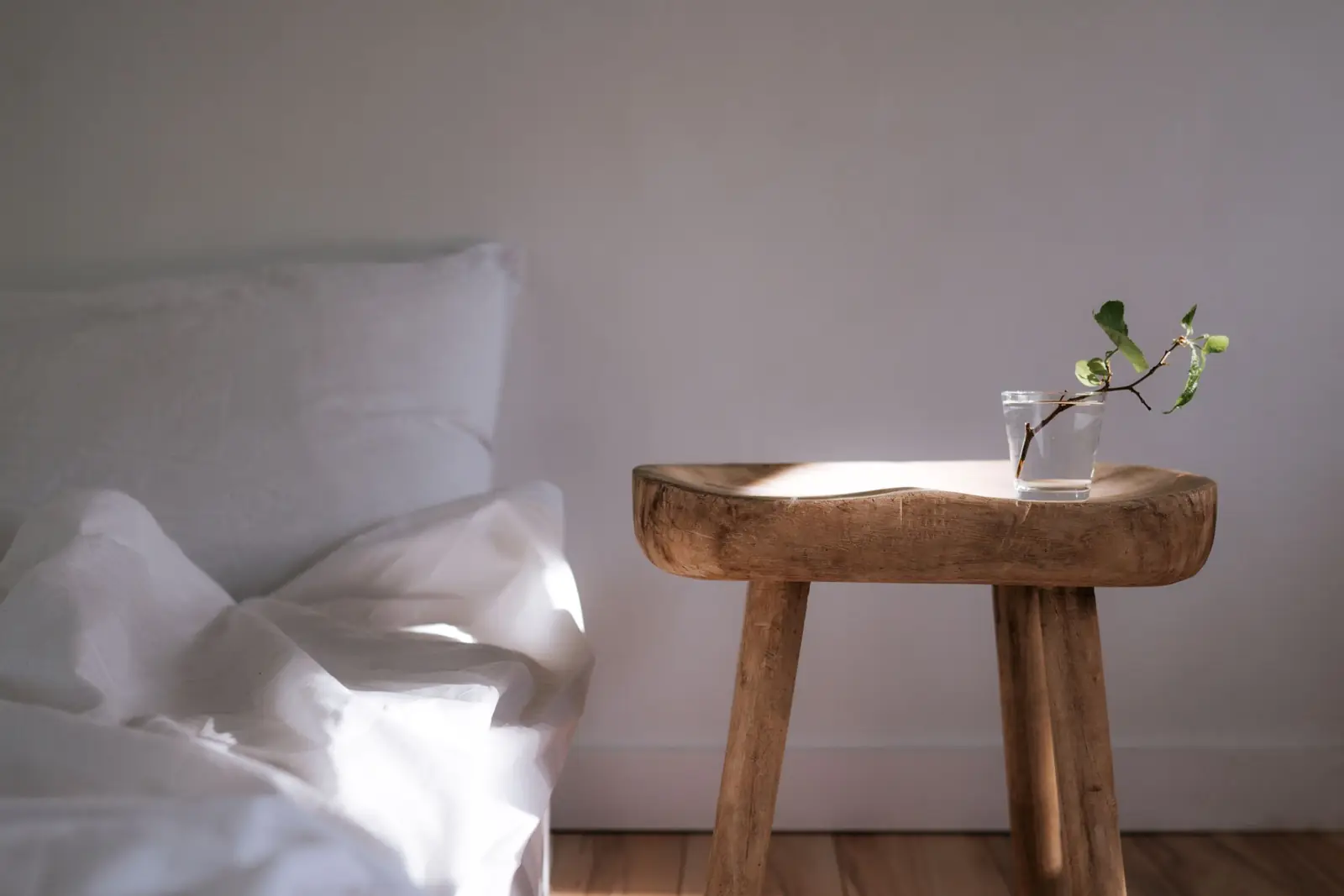 Banco de madeira ao lado da cama com um copo d'água e um raminho verde, iluminado pelo sol da manhã