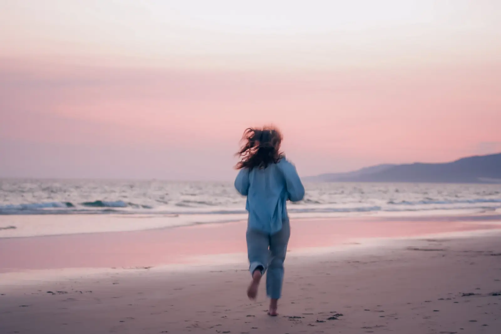 Mulher de costas, descalça, corre na areia de uma praia ao entardecer, com o céu rosado e o mar ao fundo