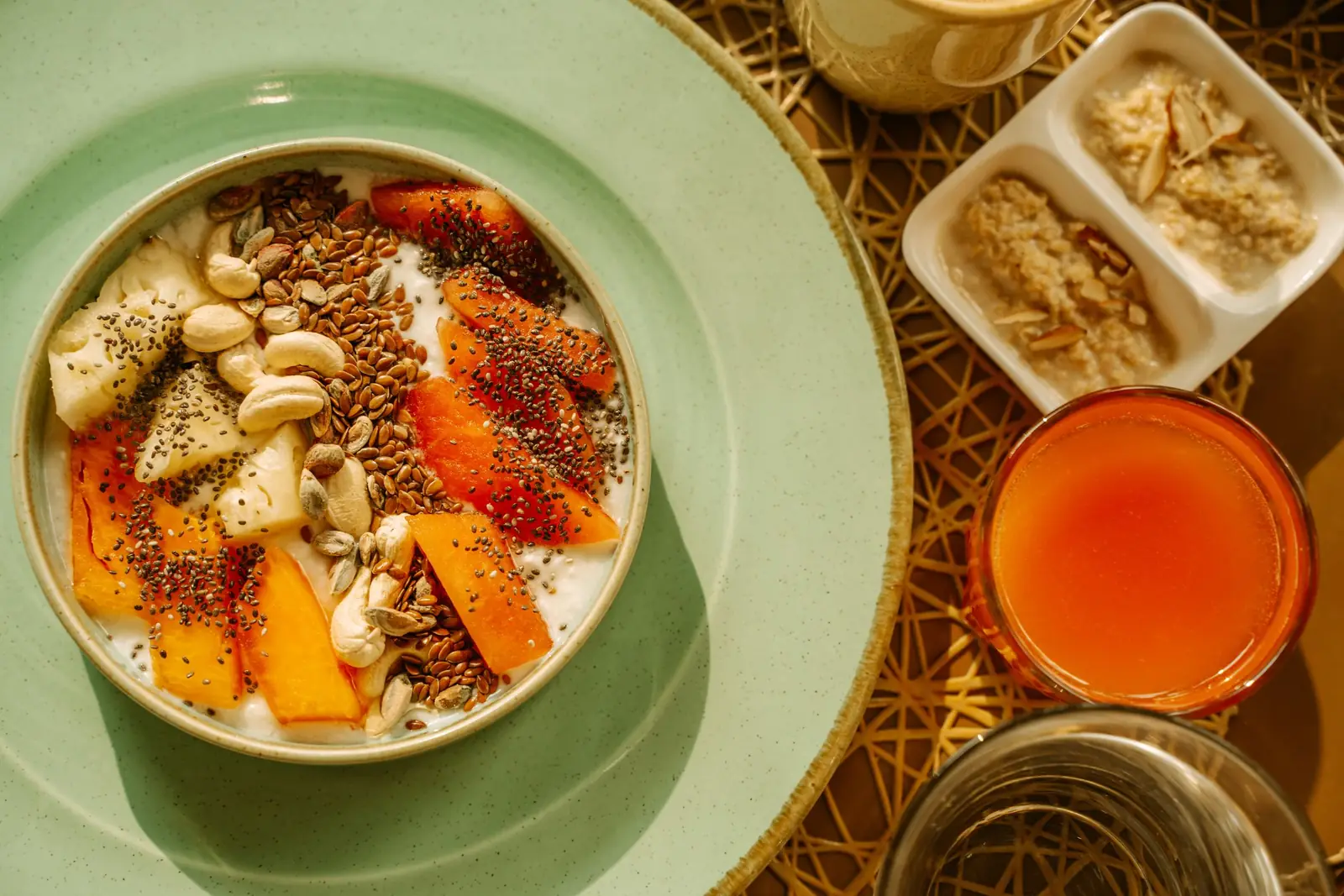 Tigela com iogurte, mamão, banana, castanhas e sementes sobre prato verde, ao lado de um copo de suco