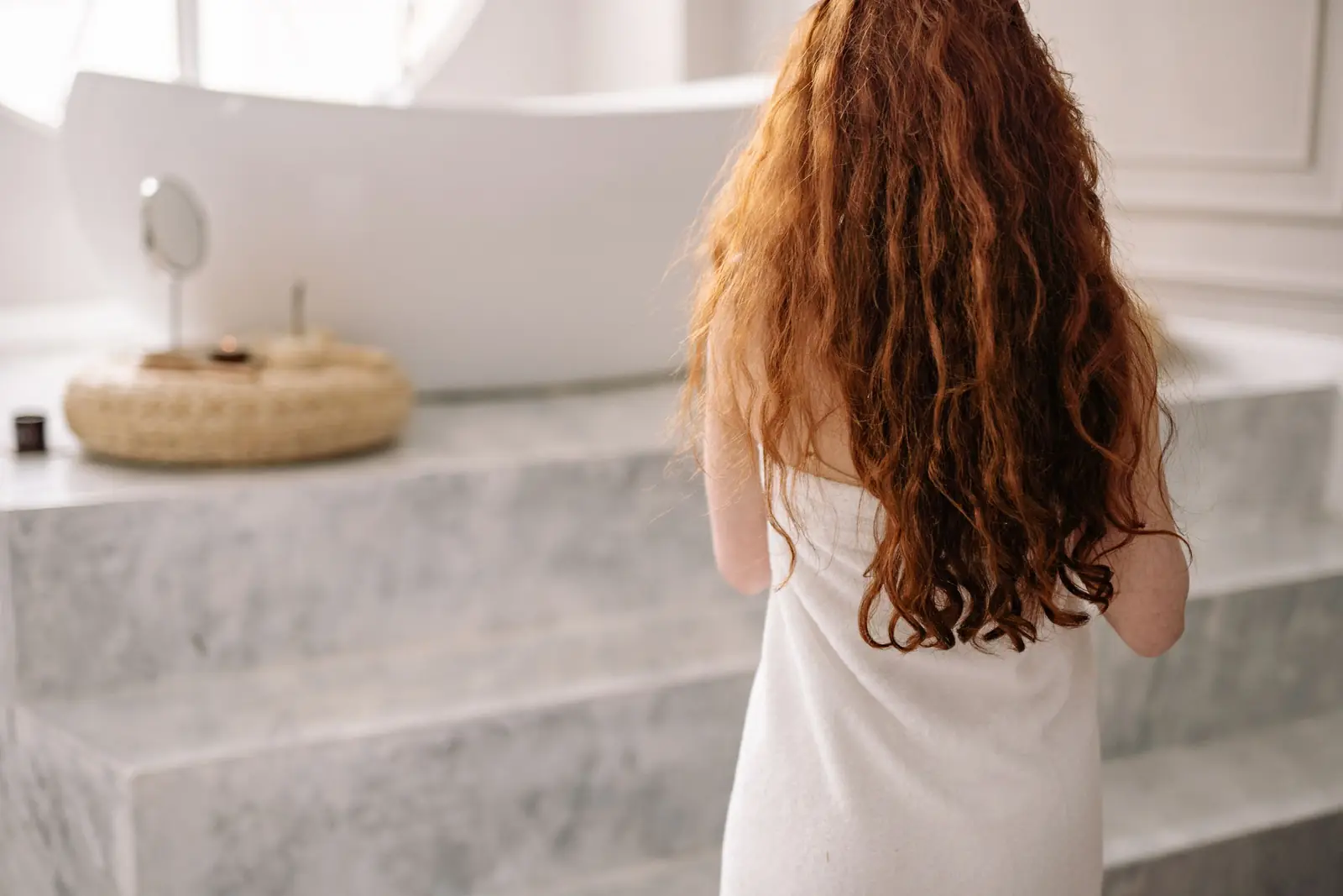 Mulher de costas, com cabelo ruivo longo e cacheado, enrolada em uma toalha no banheiro