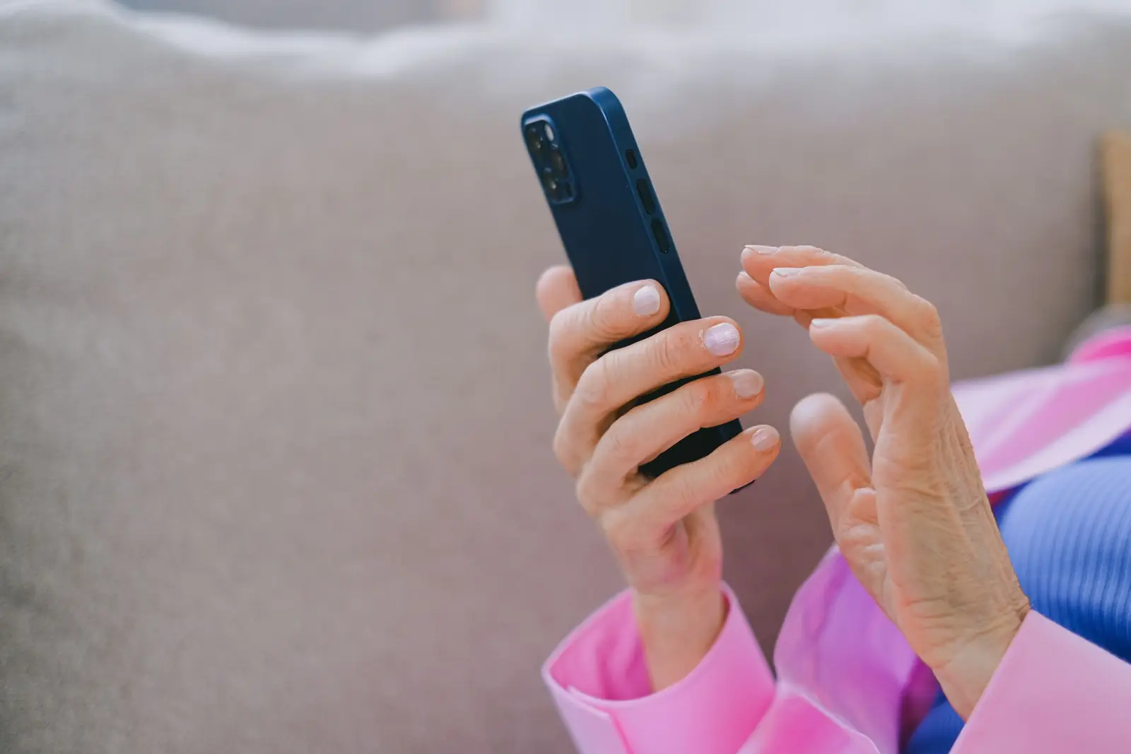 Mãos de mulher de blusa rosa segurando um celular azul, sentada em um sofá cinza