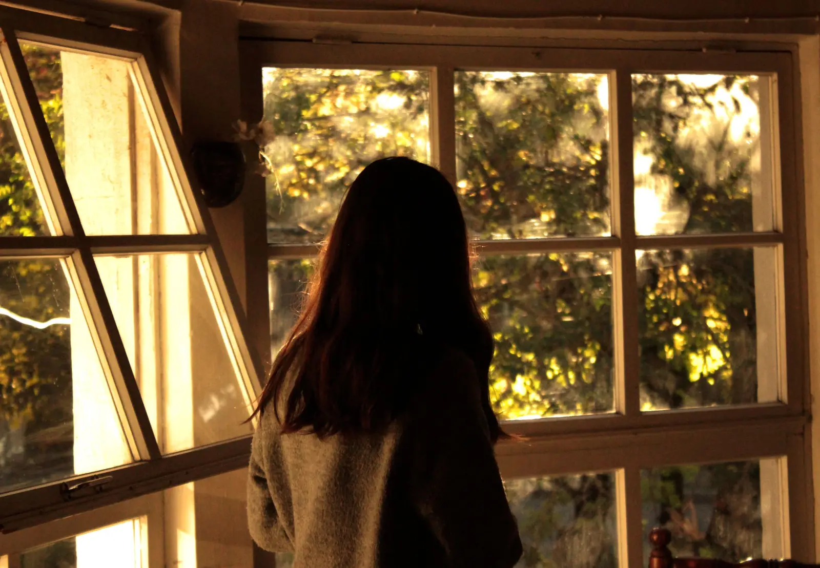 Mulher de cabelo longo, vista de costas, diante de uma janela ampla iluminada pelo sol da tarde