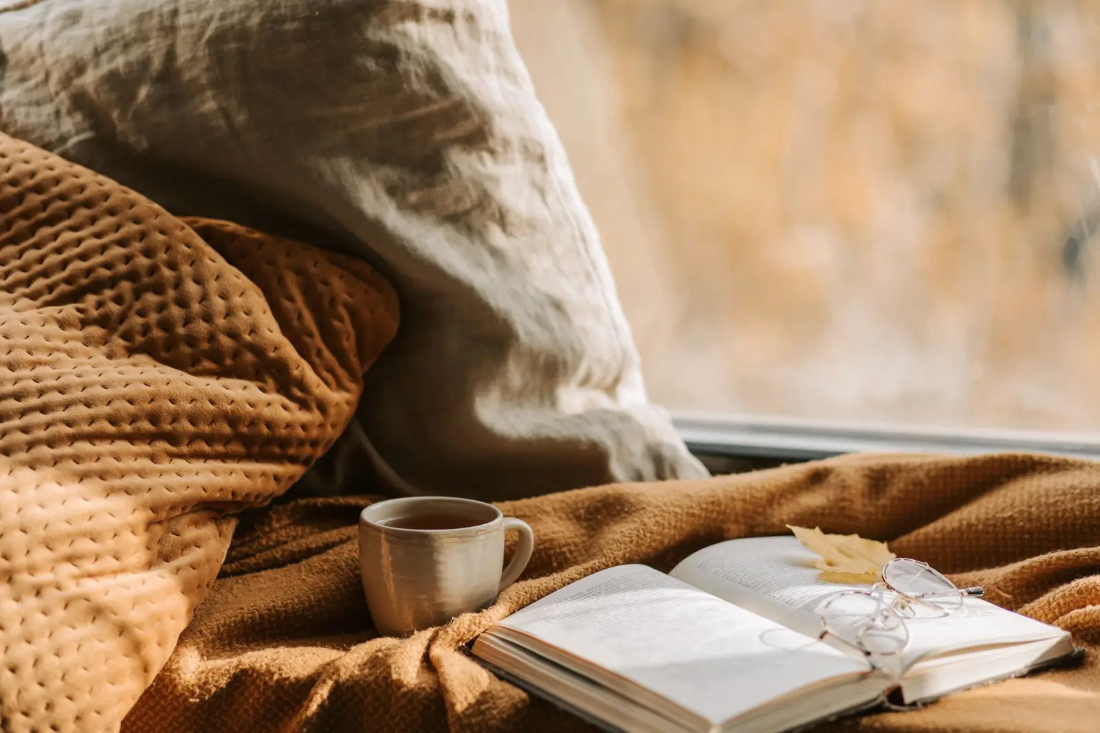 Xícara de chá e livro aberto com óculos sobre uma manta caramelo, junto à janela com luz suave