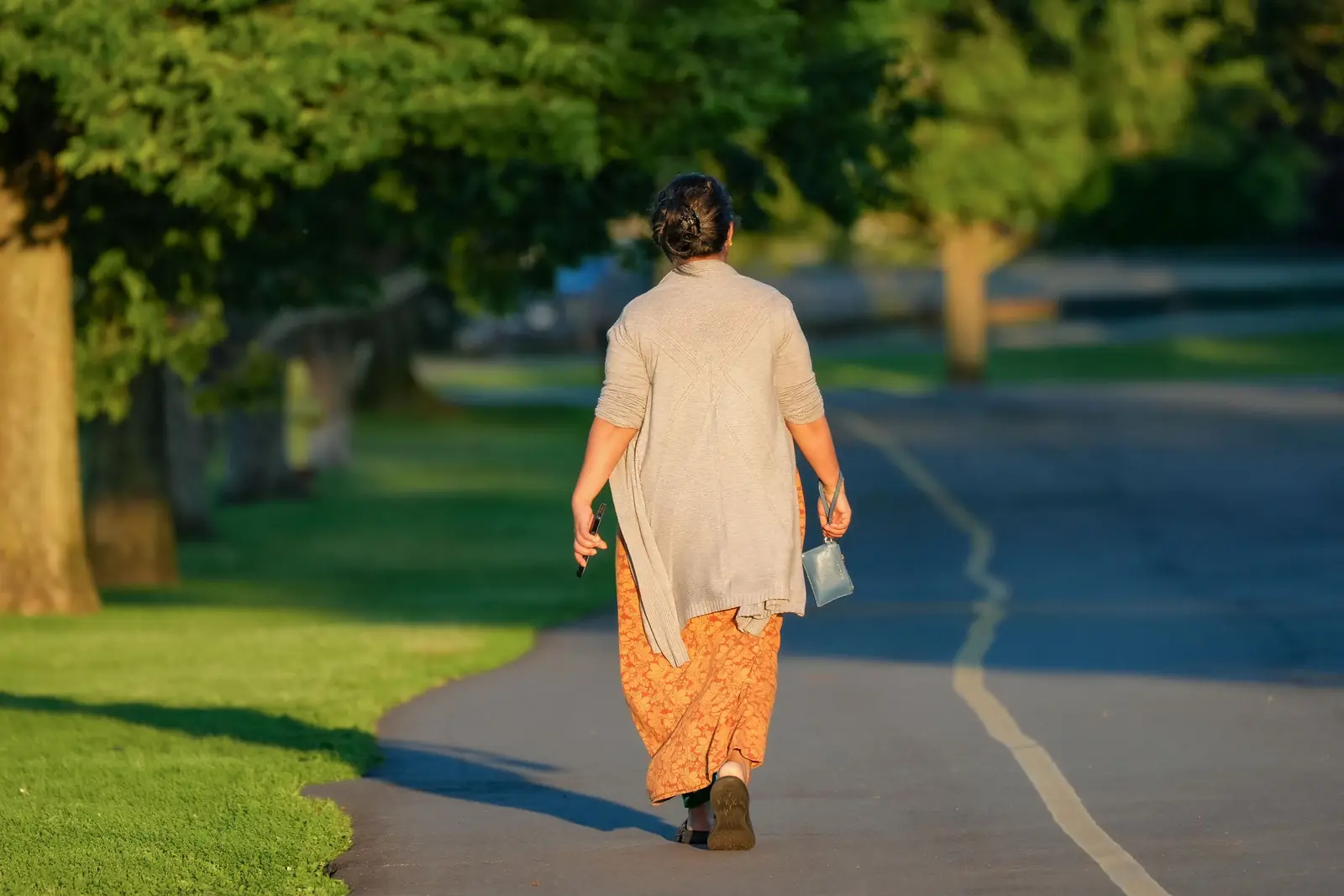 Mulher de costas caminhando por uma alameda arborizada de um parque, sob a luz dourada da manhã