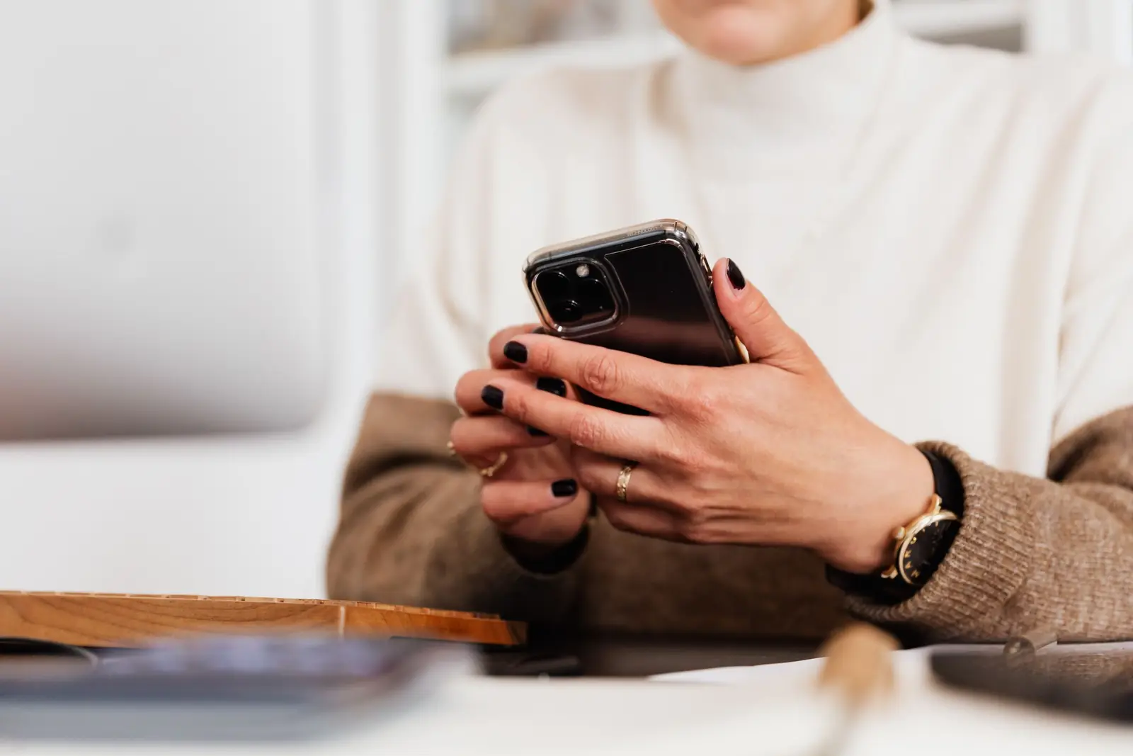 Mulher de suéter claro segura o celular com as duas mãos, sentada à mesa, com o rosto fora do quadro